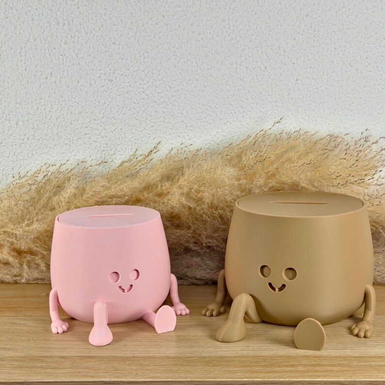 May include: Two rounded coin banks, one light pink and one tan, each featuring a simple cutout face with two circular eyes and a small curved mouth. Both containers have small, molded arms and legs extending from the sides and base.