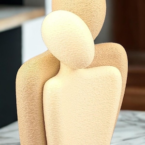 May include: A beige sculpture depicting two abstract figures in an embrace. The figures have smooth, rounded, featureless heads and torsos with a textured, grainy surface finish. The taller figure stands behind the shorter one, creating a unified form.