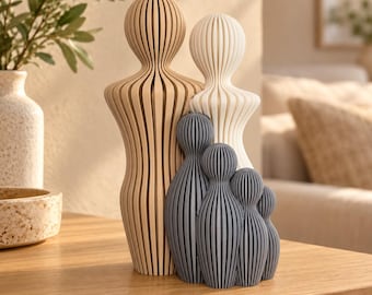 Op de afbeelding: Een set van vijf abstracte sculpturen met verticale lamellen en afgeronde hoofden. De grootste figuur is lichtbruin, de tweede is gebroken wit en de drie kleinere figuren zijn donkergrijs. Elk stuk heeft een geribbelde, gelaagde structuur.