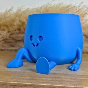 May include: A bright blue planter with a smooth matte finish, featuring a rounded body, two extended legs with feet, and a simple carved face consisting of two circular eyes and a small curved mouth, designed to sit on a flat surface.