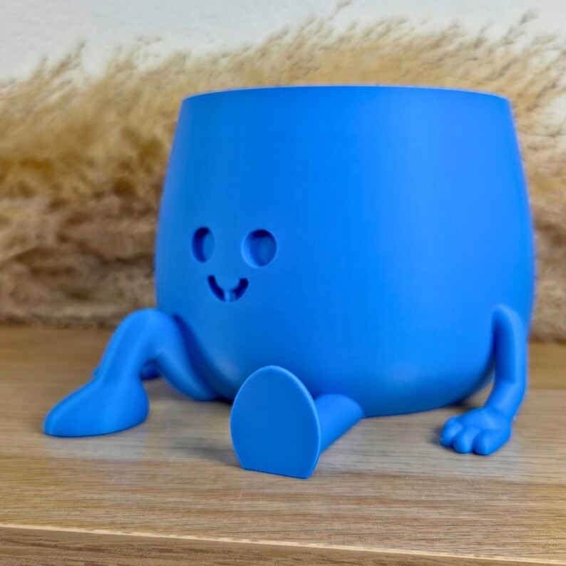 May include: A bright blue planter with a smooth matte finish, featuring a rounded body, two extended legs with feet, and a simple carved face consisting of two circular eyes and a small curved mouth, designed to sit on a flat surface.