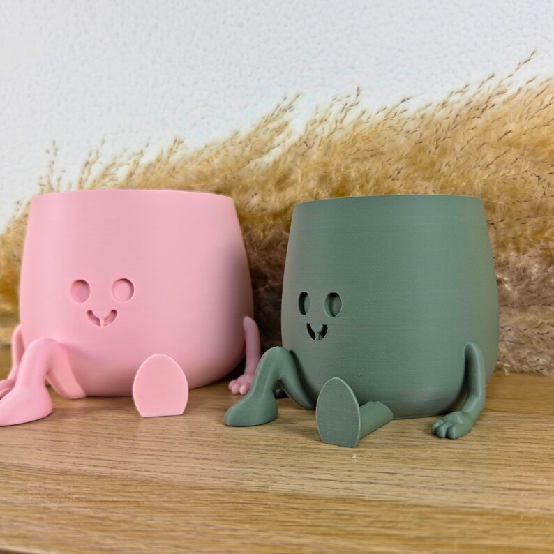 May include: Two small planters, one light pink and one sage green, each featuring a smooth matte finish, a rounded body, and a carved face with circular eyes and a curved mouth. Both planters have attached arms and legs in a seated position.