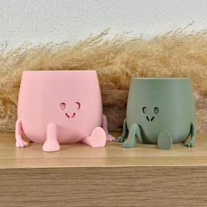 May include: Two matte planters, one light pink and one sage green, each featuring a smooth, rounded body with a simple cutout face consisting of two circular eyes and a small curved mouth, plus two small arms and two legs extending outward.