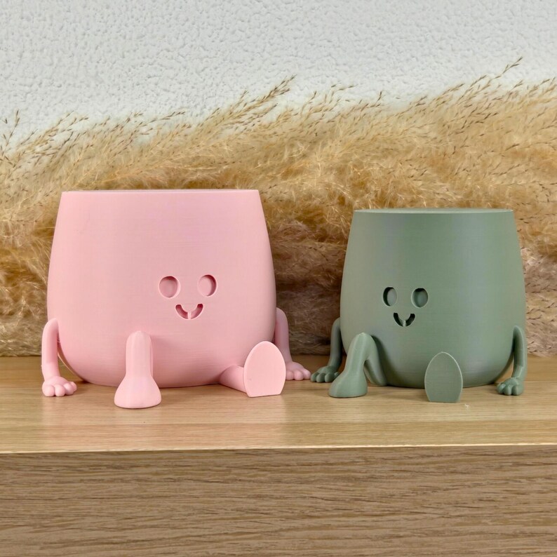 May include: Two matte planters, one light pink and one sage green, each featuring a smooth, rounded body with a simple cutout face consisting of two circular eyes and a small curved mouth, plus two small arms and two legs extending outward.