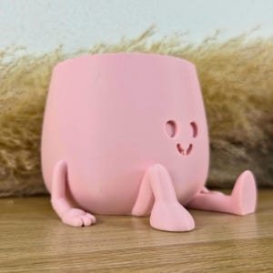 May include: A light pink planter shaped like a seated character with a smooth matte finish. The planter features a simple face with two circular eyes and a small curved mouth, along with two arms and two legs extending from the base.