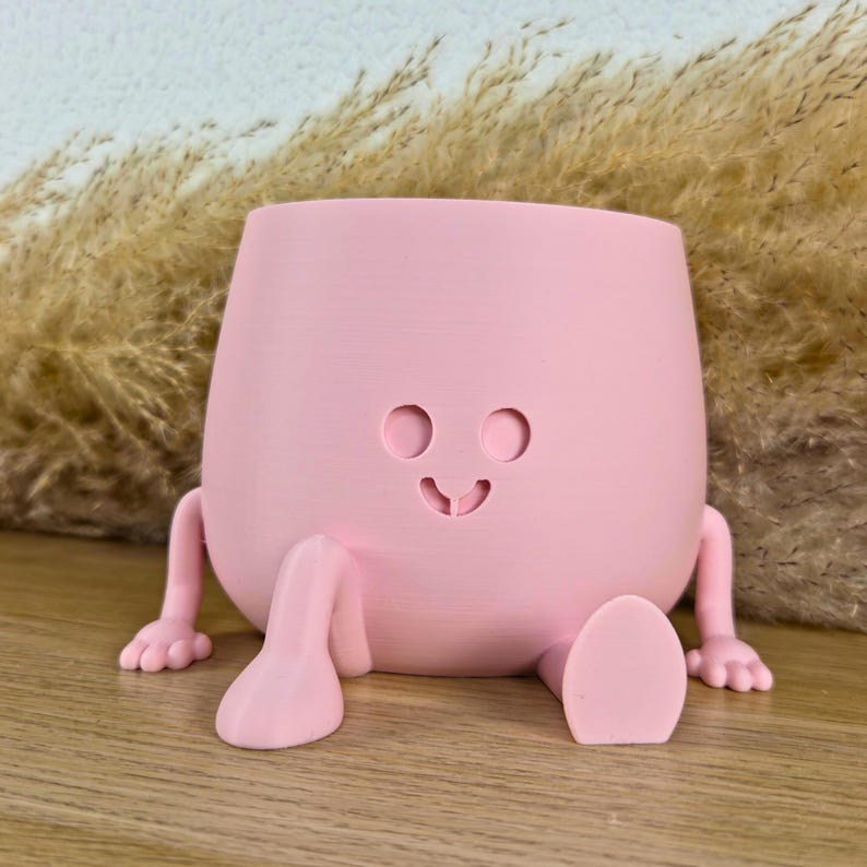 May include: A light pink planter with a smooth matte finish, featuring a rounded body, two small arms with visible fingers, and two legs with feet. The front of the planter displays a simple carved face with two circular eyes and a small mouth.