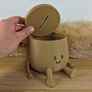 May include: A tan-colored, rounded container with a removable lid featuring a coin slot. The container has a small, simple face with two circular eyes and a curved mouth, along with two arms and two legs extending from the base in a seated position.