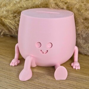 May include: A light pink, rounded container with a flat top slot, featuring two small arms and two legs extending from the base. The front surface displays a simple, engraved face with two circular eyes and a small, curved mouth.