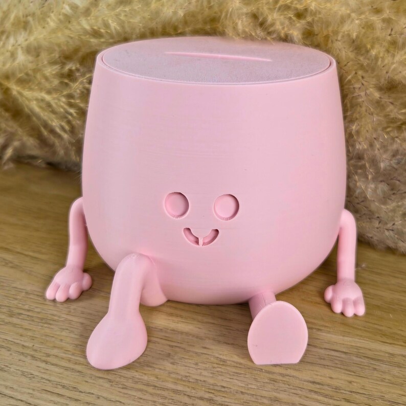May include: A light pink, rounded container with a flat top slot, featuring two small arms and two legs extending from the base. The front surface displays a simple, engraved face with two circular eyes and a small, curved mouth.