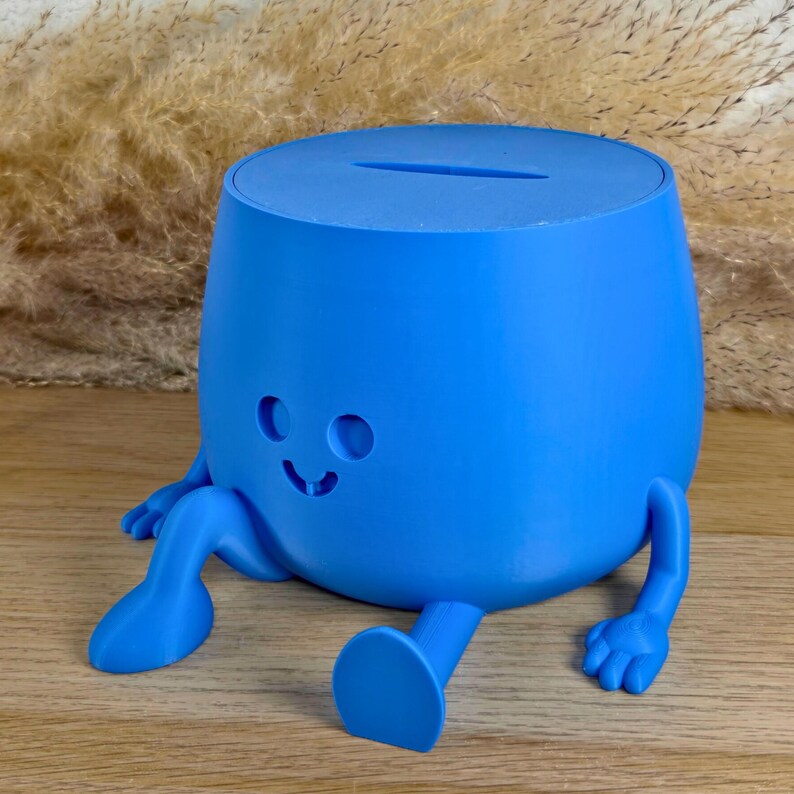 May include: A bright blue, rounded piggy bank featuring a smooth matte finish, a coin slot on the top, and a molded face with two circular eyes and a small smiling mouth. The container has two extended arms and two legs positioned in a seated pose.