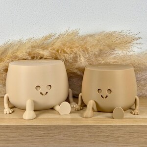 May include: Two beige, rounded containers with slots on top, each featuring a simple cutout face with circular eyes and a curved mouth. Both containers have small, articulated arms and legs extending from their sides, resting in a seated position.