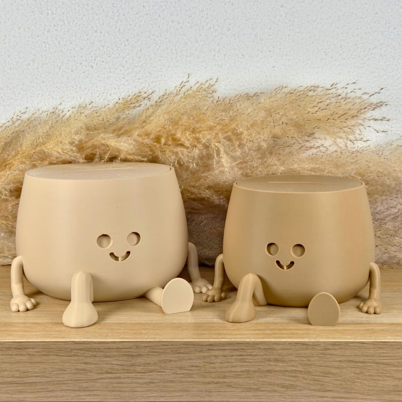 May include: Two beige, rounded containers with slots on top, each featuring a simple cutout face with circular eyes and a curved mouth. Both containers have small, articulated arms and legs extending from their sides, resting in a seated position.