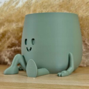 May include: A sage green planter shaped like a seated character with a smooth, matte finish. The planter features two cutout eyes and a small, simple mouth, with two extended legs and two arms resting on the surface in a relaxed pose.