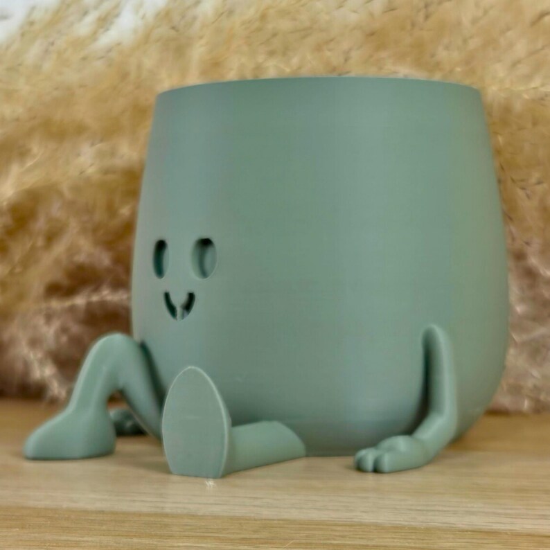 May include: A sage green planter shaped like a seated character with a smooth, matte finish. The planter features two cutout eyes and a small, simple mouth, with two extended legs and two arms resting on the surface in a relaxed pose.
