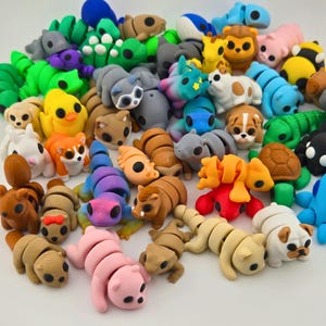 Peut inclure: Une grande collection de figurines d'animaux articulées imprimées en 3D de différentes couleurs et espèces, incluant un poussin jaune, un cochon rose, une tortue brune, une baleine bleue, un lézard vert et plusieurs chiens, tous à corps segmentés.