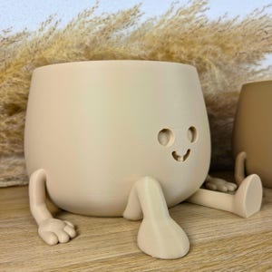May include: A light beige, rounded planter with a smooth finish, featuring two small circular eyes and a curved mouth cutout. The planter has two extended arms with hand-like ends and two legs with foot-like ends, giving it a seated appearance.