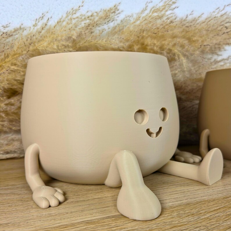 May include: A light beige, rounded planter with a smooth finish, featuring two small circular eyes and a curved mouth cutout. The planter has two extended arms with hand-like ends and two legs with foot-like ends, giving it a seated appearance.