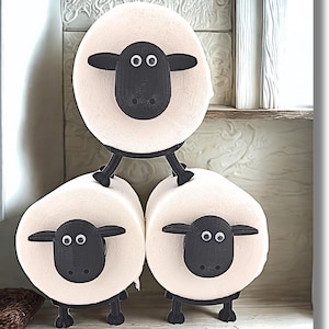 May include: A toilet paper holder shaped like three sheep, featuring black plastic heads with googly eyes and black legs attached to three rolls of white toilet paper. The sheep are arranged in a pyramid formation, with one sheep on top and two on the bottom, creating a functional storage display for bathroom tissue.