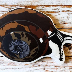 May include: A decorative pillow shaped like a snail. The pillow features a brown and tan shell design with a white body. The shell has a spiral pattern with shades of brown, tan, and black. The snail's body is white with black and white details.