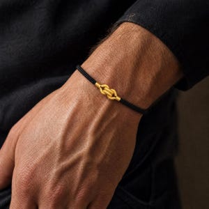May include: A black cord bracelet featuring a central gold-colored knot ornament. The bracelet consists of a thin, textured black cord that encircles the wrist, connected by a metallic gold-toned knot piece with a matte, granular surface texture.
