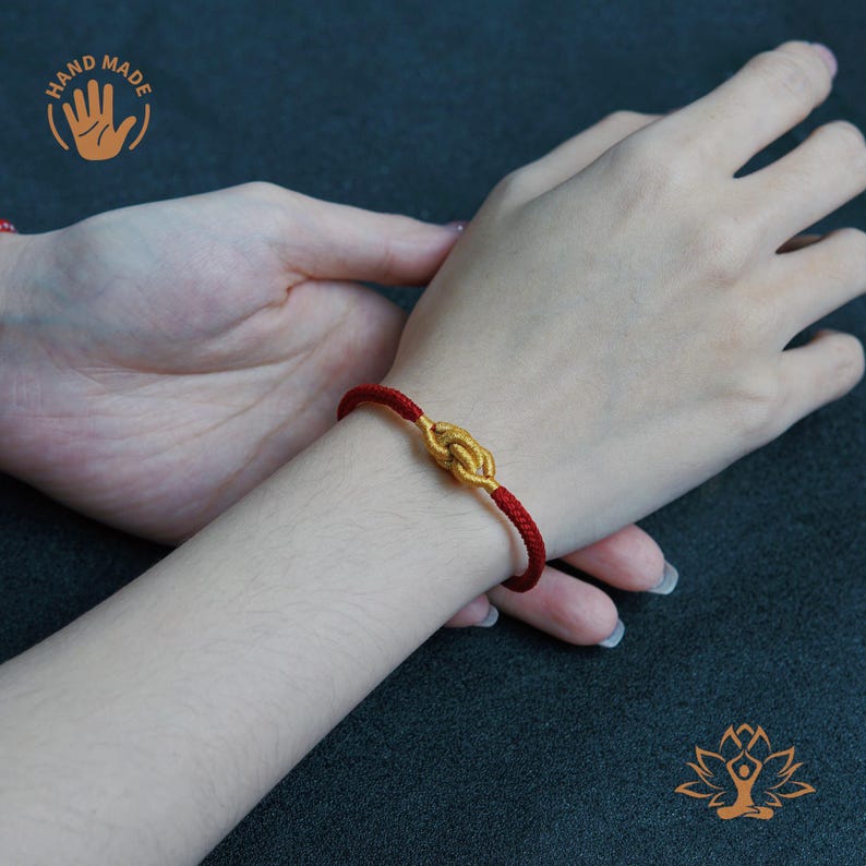 Puede incluir: Se muestra una pulsera roja y dorada en una mu&ntilde;eca. La pulsera presenta un dise&ntilde;o de nudo dorado en el centro y un cord&oacute;n rojo. La imagen tambi&eacute;n muestra una mano con las palabras "Hand Made" en un c&iacute;rculo.