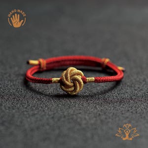 May include: Handmade red and gold bracelet featuring a central gold knot design. The bracelet is made of woven cord and has gold accents. The image shows the bracelet on a dark gray surface.