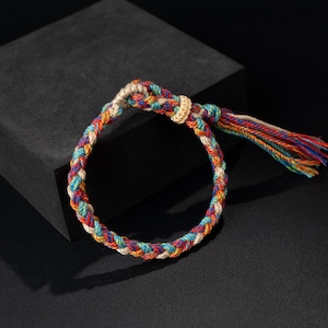 以下が含まれることがあります： カラフルなデザインの編み込みブレスレット。赤、オレンジ、黄色、緑、青、紫など、虹色の色が混ざっています。ブレスレットの端にはタッセルがあり、金色の結び目があります。
