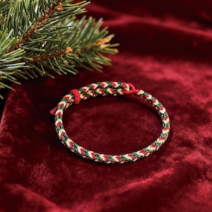 May include: A braided bracelet with red, green, and white threads. The bracelet is circular and has a woven pattern. A sprig of evergreen is in the background, and the bracelet is set against a red velvet backdrop.