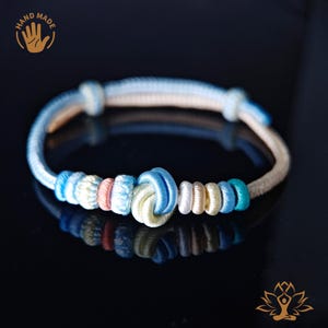 Op de afbeelding: Handgemaakte armband met een centraal knoopontwerp met blauwe, witte en roze accenten. De armband heeft een lichtblauw en beige koord. De afbeelding bevat de tekst "HAND MADE" en een handafbeelding.