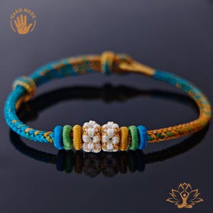 Könnte beinhalten: Handgefertigtes Armband mit einem gewebten Design in Blau-, Gelb- und Goldtönen. Das Armband hat weiße und goldene dekorative Knoten und Akzente in Grün und Blau. Das Armband wird auf einer dunklen Oberfläche präsentiert.