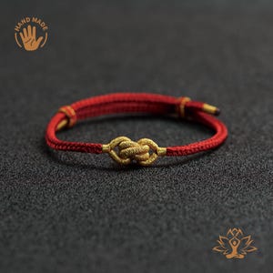 Op de afbeelding: Handgemaakte rood-gouden armband met een geweven knoopontwerp. De armband is gemaakt van rood koord met gouden accenten en een gouden knoop in het midden. De afbeelding bevat een "HAND MADE" logo.