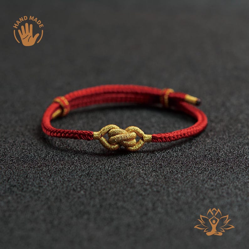 Puede incluir: Pulsera roja y dorada hecha a mano con un dise&ntilde;o de nudo tejido. La pulsera est&aacute; hecha de cord&oacute;n rojo con detalles dorados y un nudo dorado en el centro. La imagen incluye un logotipo "HAND MADE".