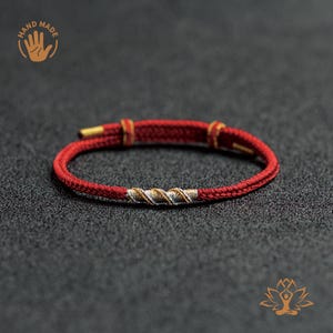 May include: A handmade red bracelet with gold detailing and a silver and gold twisted central section. The bracelet is displayed on a dark grey surface. The image also includes the text "HAND MADE".