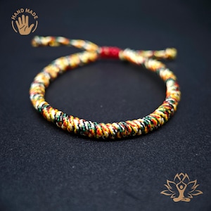 Può includere: Braccialetto fatto a mano con un design intrecciato in rosso, giallo, verde e nero. Il braccialetto è legato con un nodo rosso. Lo sfondo è nero. Il braccialetto è un gioiello.