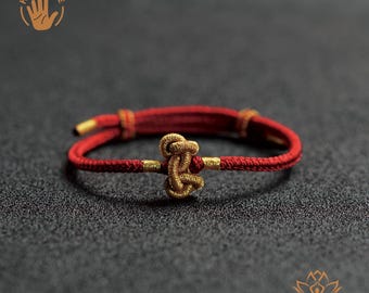 May include: A handmade red bracelet with a gold knot design. The bracelet features gold accents and a sliding knot closure. The image includes the text "HAND MADE" and a hand graphic.