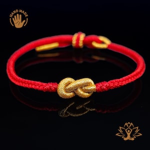 Puede incluir: Una pulsera de cordón rojo con un nudo dorado en el centro. La pulsera tiene detalles dorados y está sobre un fondo negro. La imagen también incluye un logotipo de "HAND MADE".