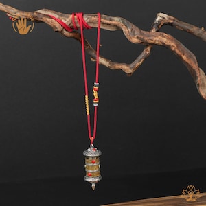 Könnte beinhalten: Eine silber-goldene Anhänger-Halskette mit roter Kordel. Der Anhänger hat eine zylindrische Form mit roten und silbernen Akzenten. Die Halskette wird auf einem braunen Ast vor dunklem Hintergrund präsentiert.