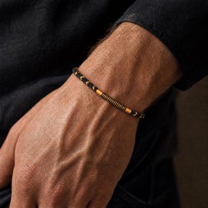 Op de afbeelding: Een dunne armband bestaande uit een zwart gevlochten koord, geaccentueerd met een centrale goudkleurige metalen cilinder met een geribbelde textuur. Het koord is geweven met subtiele goudkleurige draden, wat een contrast vormt met het zwarte materiaal.