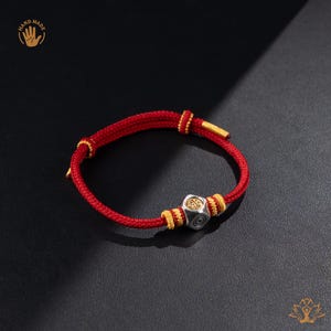May include: Red cord bracelet with gold accents and a silver, hexagonal charm. The charm has an engraved design. The bracelet is handmade.