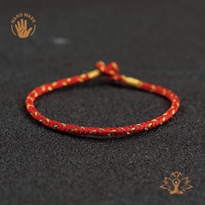 Puede incluir: Una pulsera roja y dorada hecha a mano. La pulsera está hecha de un cordón rojo trenzado con detalles dorados. La pulsera es circular y parece estar atada en los extremos. Las palabras "HAND MADE" están en la esquina superior izquierda.