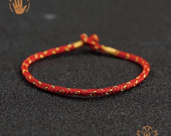 May include: A thin, braided bracelet made of vibrant red cord interwoven with metallic gold thread. The bracelet features a simple adjustable knot closure at the top. A circular logo with a hand icon and the text HAND MADE is in the top left corner.