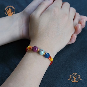 May include: Handmade bracelet featuring a series of colorful beads in shades of red, purple, blue, and turquoise. The bracelet is strung on a red and yellow cord. The image includes the text "HAND MADE" and a hand symbol.