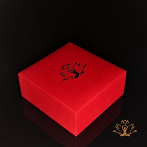 May include: A red square box with a textured surface. A black lotus flower design is on the top of the box. The box is on a black surface.