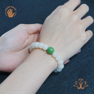May include: A handmade bracelet with white, spherical beads and a single green bead. The bracelet is shown on a wrist, highlighting its design and colour contrast. The image includes a "HAND MADE" logo.