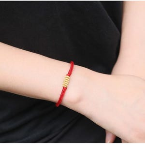 May include: A red cord bracelet featuring a central gold-colored cylindrical bead with a ribbed, textured surface. The bracelet is constructed from a thin, braided red string that wraps around the wrist, secured by the metallic gold-toned accent.