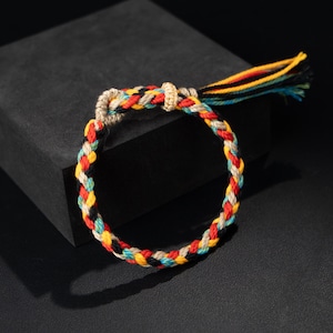 Può includere: Braccialetto intrecciato con una vivace miscela di colori: rosso, giallo, nero, blu e beige. Il braccialetto ha un design intrecciato con un nappa all'estremità. Un accessorio alla moda.