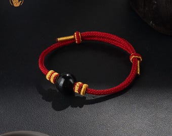 May include: Red cord bracelet with a black bead and gold-colored accents. The bracelet is handmade and features a sliding knot closure. The design is simple and elegant, suitable for everyday wear.