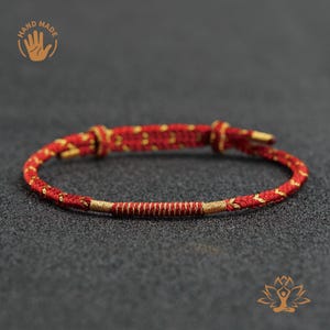 May include: Red and gold handmade bracelet. The bracelet is made of a red cord with gold accents and a gold clasp. The text "HAND MADE" is visible in the upper left corner.