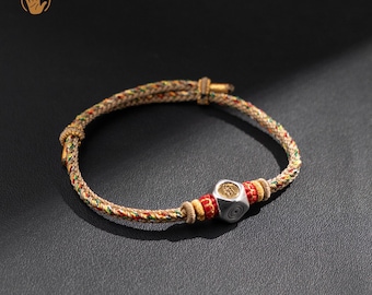 May include: A handcrafted bracelet featuring a woven cord in shades of beige, red, yellow, and green. The bracelet has a silver-colored, cube-shaped bead with an engraved design and red and yellow accent beads.