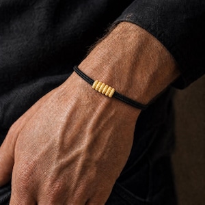 Könnte beinhalten: Ein schwarzes Kordelarmband mit fünf kleinen, zylindrischen, goldfarbenen Perlen, die eine texturierte, gerippte Oberfläche aufweisen und in einer Reihe in der Mitte des Bandes angeordnet sind. Das Armband hat einen einfachen, verstellbaren Knoten.